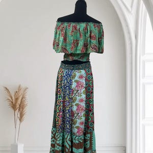 May include: A two-piece bohemian outfit on a mannequin. The off-the-shoulder crop top is green with a red and gold paisley pattern. The long skirt is a patchwork of floral and geometric patterns in shades of blue, green, and brown.