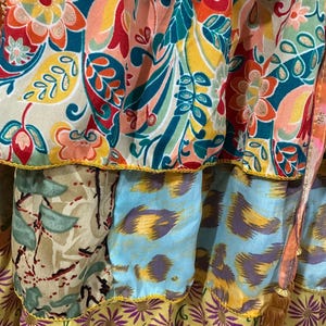 May include: Close-up of a tiered skirt with vibrant floral and abstract patterns. The skirt features a mix of colors including teal, orange, yellow, and purple. The edges are trimmed with yellow and gold accents, and a brown tassel hangs from the side.