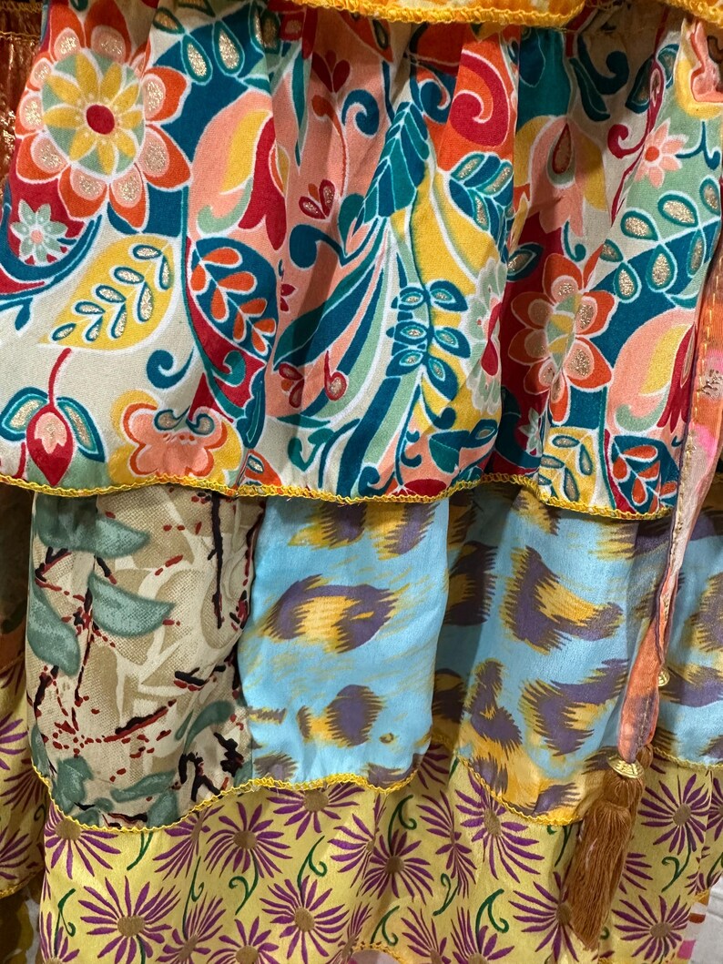 May include: Close-up of a tiered skirt with vibrant floral and abstract patterns. The skirt features a mix of colors including teal, orange, yellow, and purple. The edges are trimmed with yellow and gold accents, and a brown tassel hangs from the side.