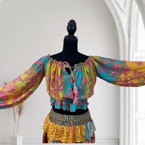 May include: A colorful bohemian-style top and skirt set. The top features long sleeves and a tie-up neckline with pink tassels. The skirt has a layered design with various patterned fabrics in yellow, blue, and pink. The outfit is displayed on a black mannequin.