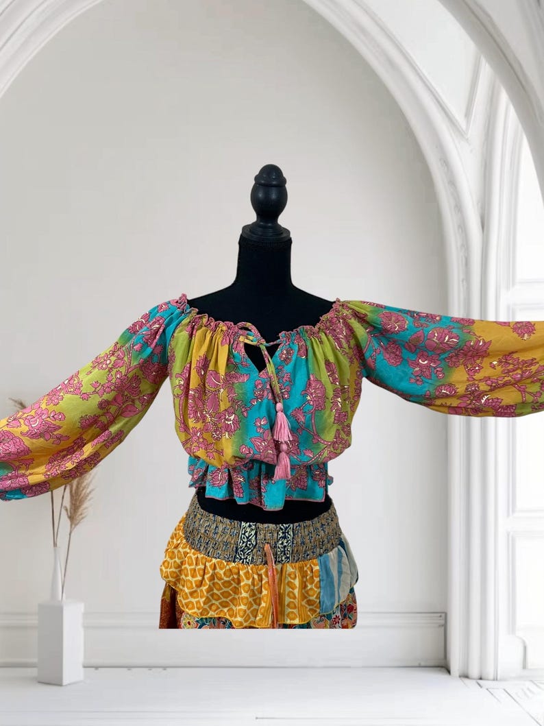 May include: A colorful bohemian-style top and skirt set. The top features long sleeves and a tie-up neckline with pink tassels. The skirt has a layered design with various patterned fabrics in yellow, blue, and pink. The outfit is displayed on a black mannequin.