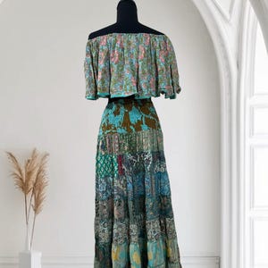 Op de afbeelding: Een tweedelige bohemien jurk op een zwarte mannequin. De off-the-shoulder top heeft een bloemenpatroon in roze, blauw en groen. De lange rok is een patchwork van verschillende stoffen met patronen in blauw, groen en bruin.