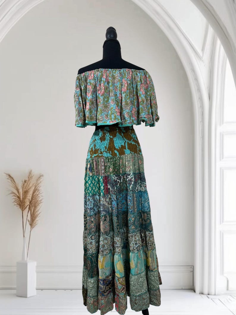 Op de afbeelding: Een tweedelige bohemien jurk op een zwarte mannequin. De off-the-shoulder top heeft een bloemenpatroon in roze, blauw en groen. De lange rok is een patchwork van verschillende stoffen met patronen in blauw, groen en bruin.