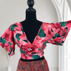 May include: A floral wrap top with a V-neck and short sleeves. The top features a pink and red floral pattern with green leaves. The top is paired with a patterned skirt with a black waistband. The outfit is displayed on a black mannequin.