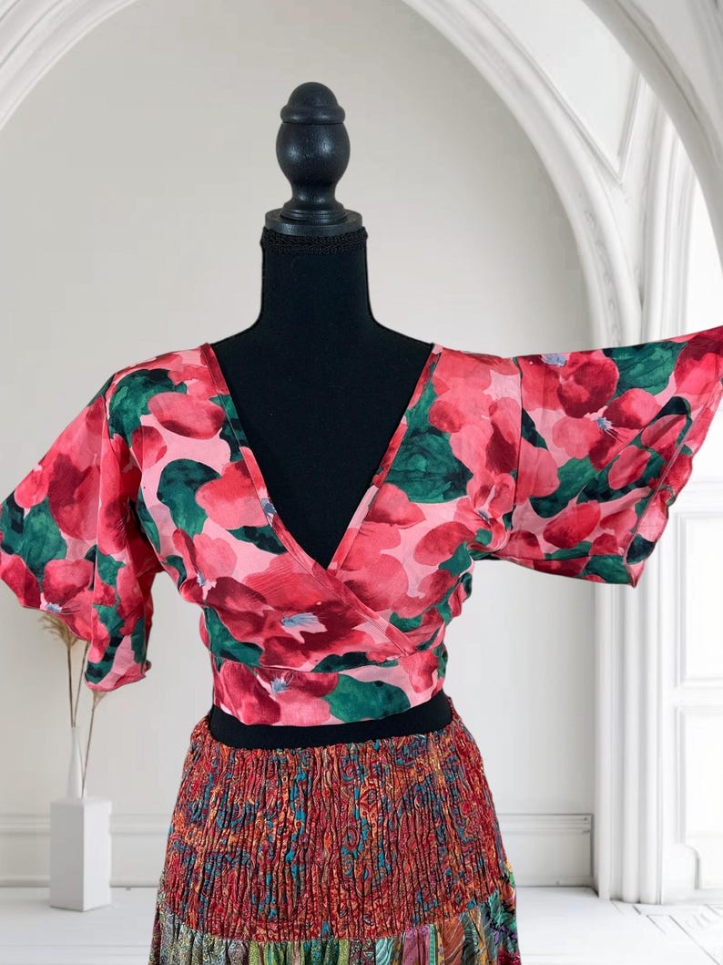 May include: A floral wrap top with a V-neck and short sleeves. The top features a pink and red floral pattern with green leaves. The top is paired with a patterned skirt with a black waistband. The outfit is displayed on a black mannequin.