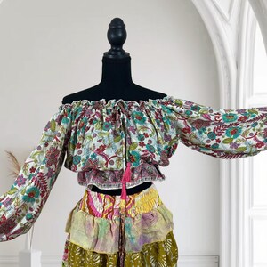 Op de afbeelding: Een off-the-shoulder top en rok met meerdere lagen. De top heeft een bloemenpatroon met rode, blauwe en groene accenten. De rok heeft meerdere lagen in verschillende kleuren en patronen, waaronder geel, roze en olijfgroen. Een roze kwast hangt aan de top.