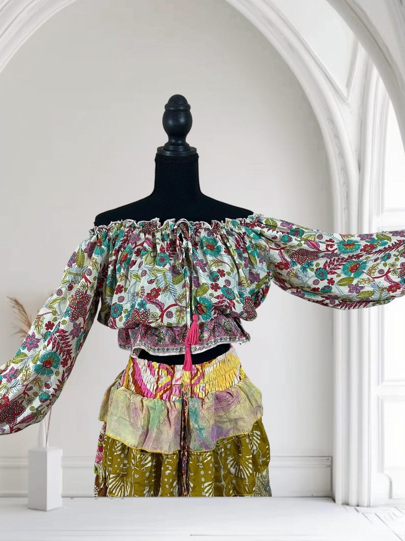 Op de afbeelding: Een off-the-shoulder top en rok met meerdere lagen. De top heeft een bloemenpatroon met rode, blauwe en groene accenten. De rok heeft meerdere lagen in verschillende kleuren en patronen, waaronder geel, roze en olijfgroen. Een roze kwast hangt aan de top.