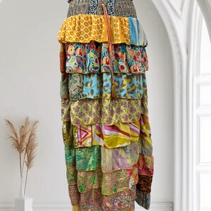 May include: A long, tiered skirt with a patchwork design. The skirt features layers of fabric in various colors and patterns, including yellow, blue, green, and paisley prints. A brown tassel hangs from the waist. The skirt is a bohemian style.