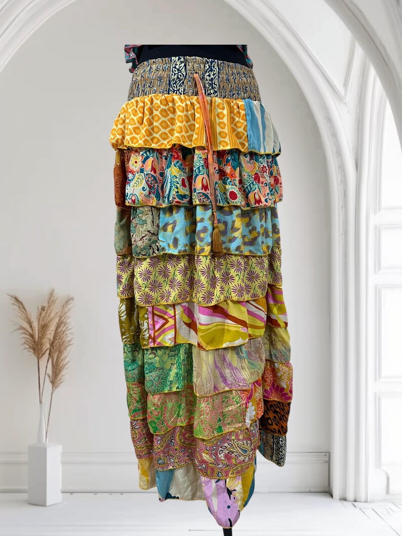 May include: A long, tiered skirt with a patchwork design. The skirt features layers of fabric in various colors and patterns, including yellow, blue, green, and paisley prints. A brown tassel hangs from the waist. The skirt is a bohemian style.