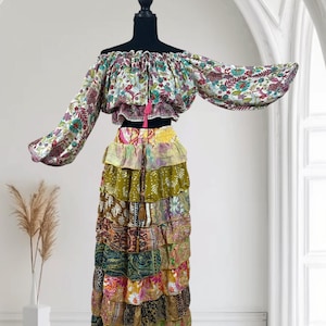 May include: A two-piece bohemian outfit on a mannequin. The off-the-shoulder top features a floral pattern with long sleeves. The skirt is a tiered maxi skirt with a patchwork design in various colours and patterns. A pink tassel hangs from the top.