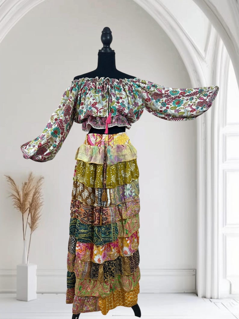 Op de afbeelding: Een tweedelige bohemien outfit op een mannequin. De off-the-shoulder top heeft een bloemenpatroon met lange mouwen. De rok is een maxi-rok met ruches en een patchwork-ontwerp in verschillende kleuren en patronen. Een roze kwast hangt van boven.