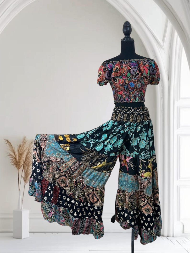 May include: A two-piece bohemian outfit on a mannequin. The off-the-shoulder crop top and wide-leg pants feature a patchwork design with floral and paisley patterns in shades of black, blue, red, and gold. The outfit is displayed against a white background.