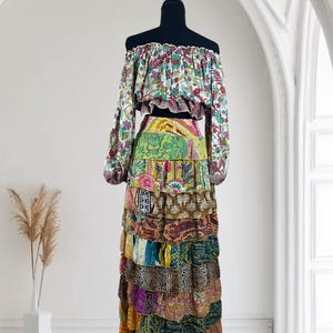 Op de afbeelding: Een tweedelige bohemien outfit op een mannequin. De off-the-shoulder top heeft een bloemenpatroon met lange mouwen. De gelaagde maxi-rok is samengesteld uit verschillende kleurrijke stoffen patches, waardoor een patchwork-ontwerp ontstaat. De outfit wordt gepresenteerd tegen een witte achtergrond.
