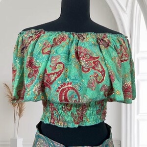 May include: An off-the-shoulder top with a green paisley pattern. The top has a ruffled neckline and a fitted, elasticated waist. The paisley design features red and gold accents on a teal background. The top is displayed on a black mannequin.