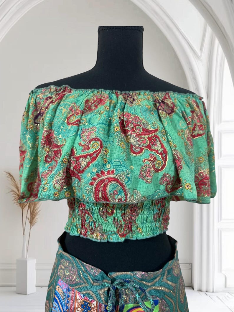 May include: An off-the-shoulder top with a green paisley pattern. The top has a ruffled neckline and a fitted, elasticated waist. The paisley design features red and gold accents on a teal background. The top is displayed on a black mannequin.
