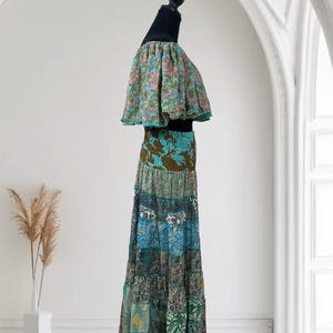 Op de afbeelding: Een tweedelige bohemien outfit op een mannequin. De top heeft een bloemenprint met roze en blauwe bloemen. De maxi-rok heeft een patchwork ontwerp in blauw, groen en bruin. De outfit wordt gepresenteerd tegen een witte achtergrond.