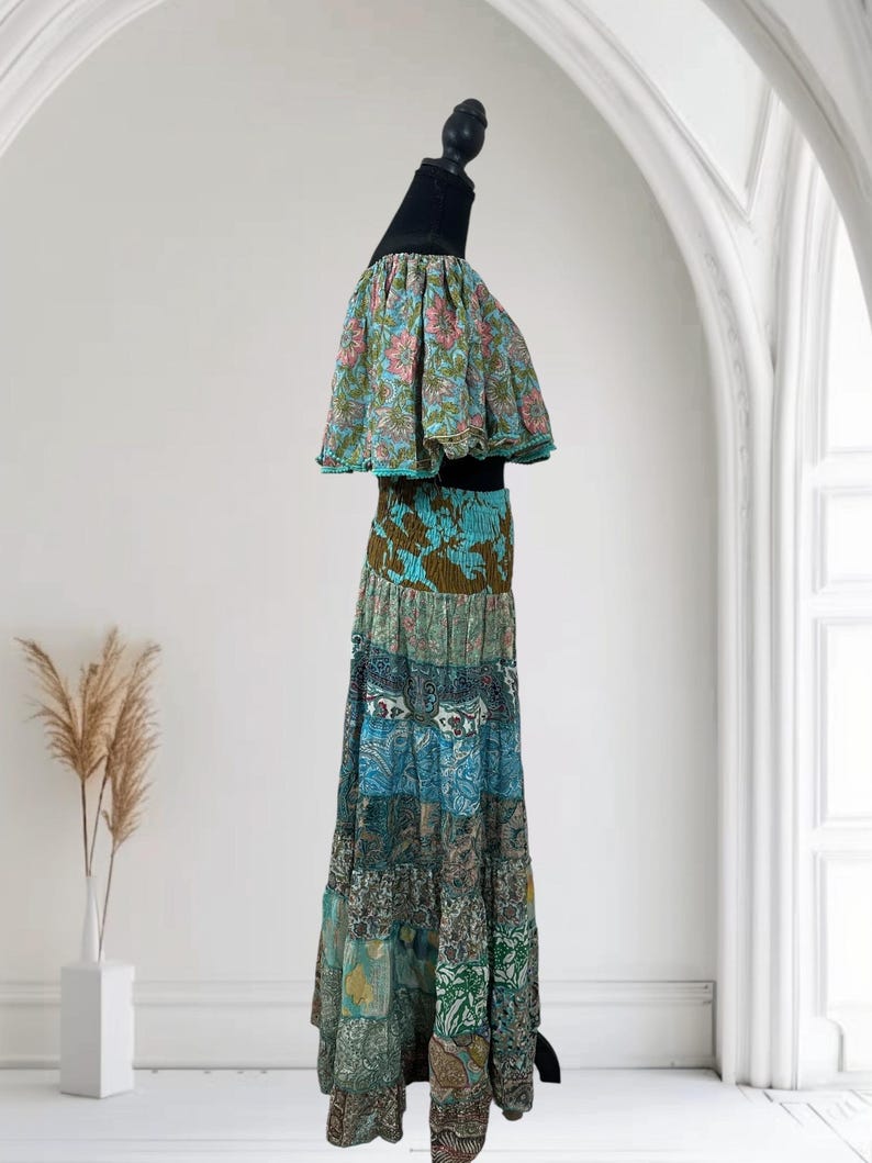 Op de afbeelding: Een tweedelige bohemien outfit op een mannequin. De top heeft een bloemenprint met roze en blauwe bloemen. De maxi-rok heeft een patchwork ontwerp in blauw, groen en bruin. De outfit wordt gepresenteerd tegen een witte achtergrond.