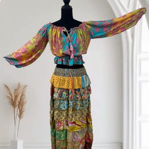 May include: A colorful bohemian two-piece outfit. The off-the-shoulder top features long sleeves and a tie-up neckline with pink tassels. The tiered skirt is made of various patterned fabrics in shades of yellow, blue, green, and pink. The outfit is displayed on a black mannequin.