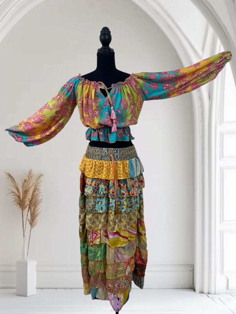 May include: A colorful bohemian two-piece outfit. The off-the-shoulder top features long sleeves and a tie-up neckline with pink tassels. The tiered skirt is made of various patterned fabrics in shades of yellow, blue, green, and pink. The outfit is displayed on a black mannequin.