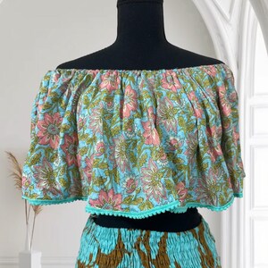 Op de afbeelding: Een tweedelige outfit met een off-the-shoulder crop top en een rok. De top is lichtblauw met een roze en groen bloemenpatroon, afgezet met turquoise pompons. De rok combineert turquoise en bruin.