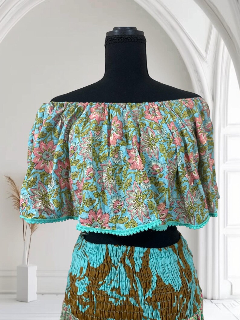 Op de afbeelding: Een tweedelige outfit met een off-the-shoulder crop top en een rok. De top is lichtblauw met een roze en groen bloemenpatroon, afgezet met turquoise pompons. De rok combineert turquoise en bruin.