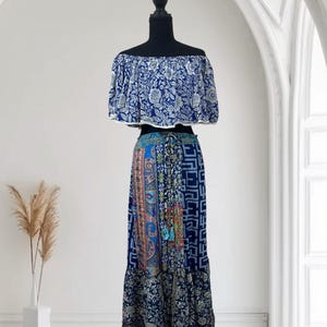 May include: A two-piece bohemian outfit on a black mannequin. The off-the-shoulder top is blue with a white floral pattern. The long skirt features a patchwork design with various blue and patterned fabrics. The skirt has a drawstring waist and a ruffled hem.