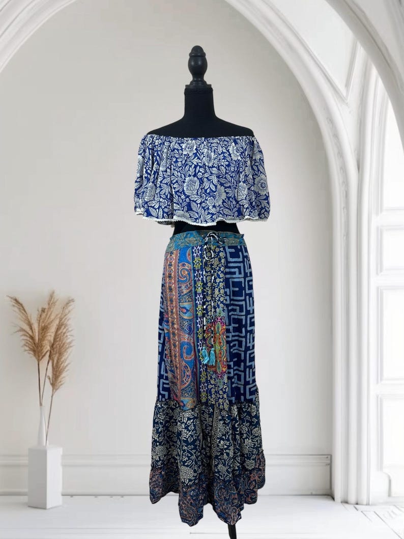 May include: A two-piece bohemian outfit on a black mannequin. The off-the-shoulder top is blue with a white floral pattern. The long skirt features a patchwork design with various blue and patterned fabrics. The skirt has a drawstring waist and a ruffled hem.