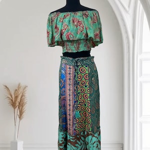 May include: A two-piece bohemian outfit featuring an off-the-shoulder crop top and a long skirt. The top is light green with a red paisley pattern. The skirt is a patchwork of green, blue, and brown fabrics with floral and geometric designs.