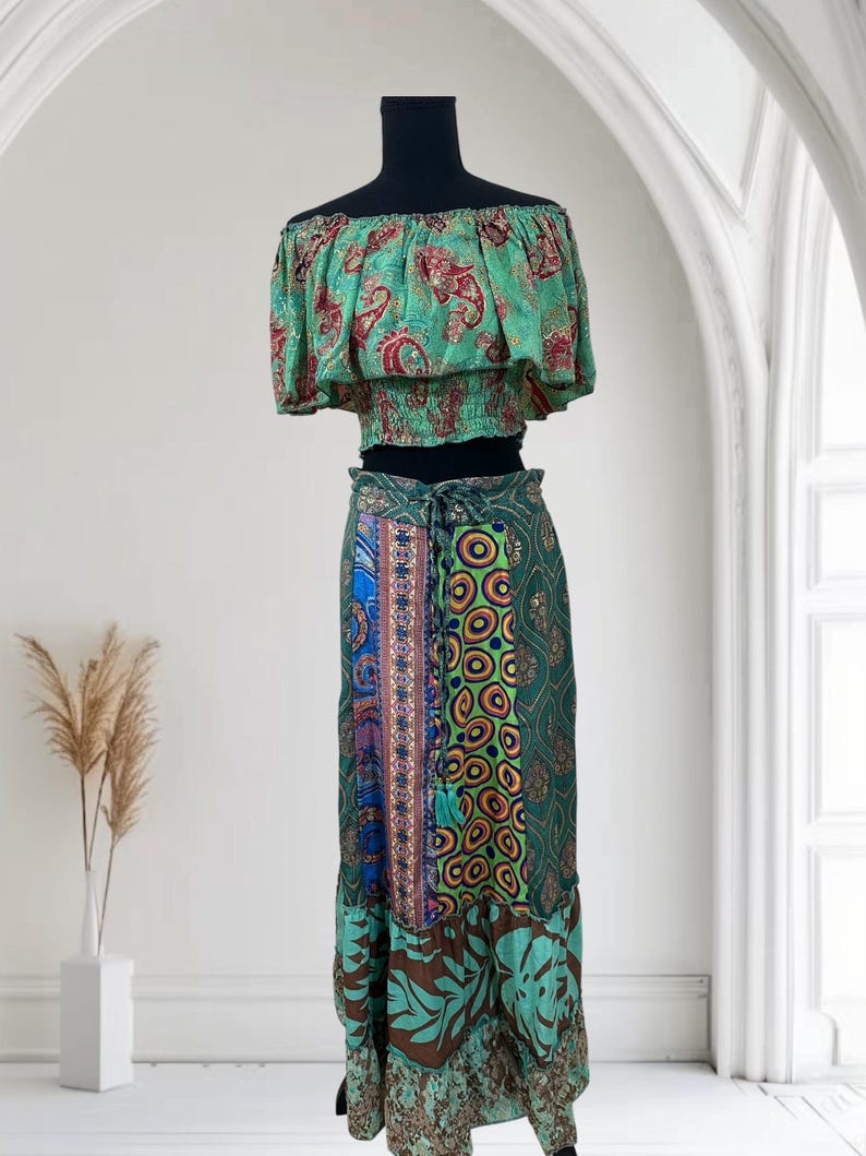May include: A two-piece bohemian outfit featuring an off-the-shoulder crop top and a long skirt. The top is light green with a red paisley pattern. The skirt is a patchwork of green, blue, and brown fabrics with floral and geometric designs.