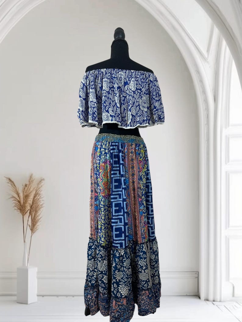 May include: A two-piece bohemian outfit on a mannequin. The off-the-shoulder top is blue with a white floral pattern. The long skirt features a patchwork design with various patterns and colors, including blue, pink, and orange. The skirt has a ruffled hem.