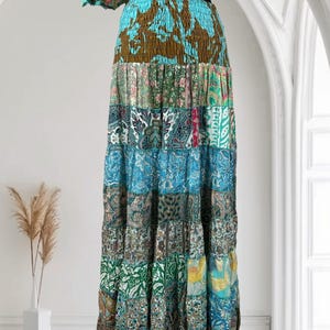 Op de afbeelding: Lange patchwork rok met een gelaagd ontwerp. De rok heeft een verscheidenheid aan stoffen met patronen in tinten blauw, groen, bruin en roze. De rok heeft een zwarte tailleband en wordt gepresenteerd tegen een witte achtergrond.