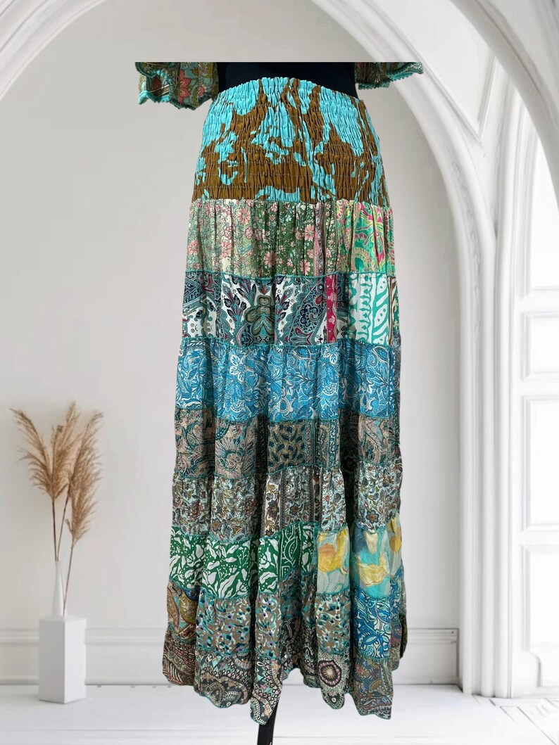 Op de afbeelding: Lange patchwork rok met een gelaagd ontwerp. De rok heeft een verscheidenheid aan stoffen met patronen in tinten blauw, groen, bruin en roze. De rok heeft een zwarte tailleband en wordt gepresenteerd tegen een witte achtergrond.