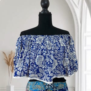 May include: Off-the-shoulder crop top with a blue and white floral pattern. The top has short sleeves and a white trim along the bottom edge. The top is displayed on a black mannequin.