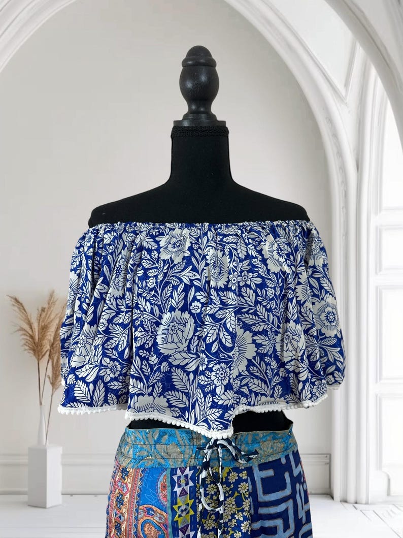 May include: Off-the-shoulder crop top with a blue and white floral pattern. The top has short sleeves and a white trim along the bottom edge. The top is displayed on a black mannequin.