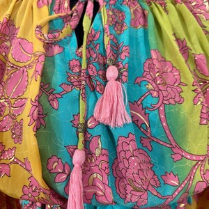 May include: A colorful top with a floral pattern in yellow, turquoise, and green. The design includes pink flowers and gold accents. Pink tassels hang from the neckline, adding a decorative touch. The top has a drawstring neckline.