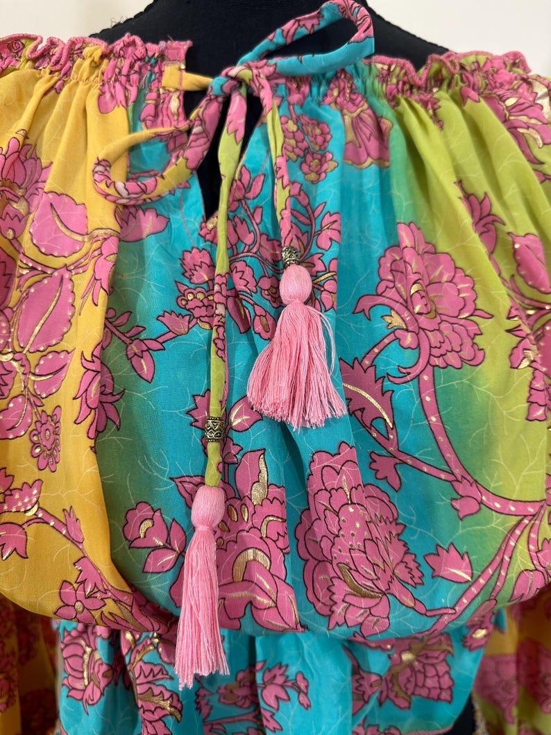 May include: A colorful top with a floral pattern in yellow, turquoise, and green. The design includes pink flowers and gold accents. Pink tassels hang from the neckline, adding a decorative touch. The top has a drawstring neckline.