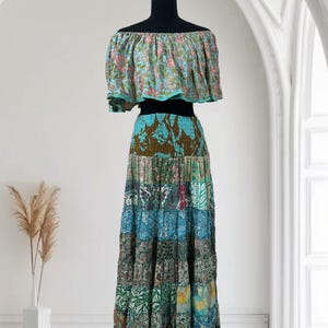 Op de afbeelding: Een tweedelige bohemien outfit op een zwarte mannequin. De off-the-shoulder top heeft een bloemenpatroon met roze en blauwe tinten. De gelaagde rok is een patchwork van verschillende stoffen met patronen in blauw, groen en bruin.