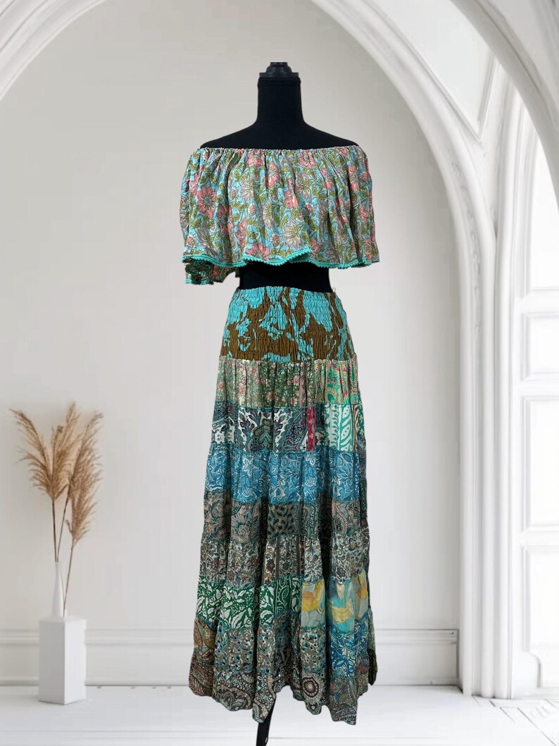 Op de afbeelding: Een tweedelige bohemien outfit op een zwarte mannequin. De off-the-shoulder top heeft een bloemenpatroon met roze en blauwe tinten. De gelaagde rok is een patchwork van verschillende stoffen met patronen in blauw, groen en bruin.