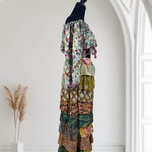 Op de afbeelding: Een kleurrijke, gelaagde maxi-jurk met een off-the-shoulder top. De top heeft een bloemenprint met gefranjerde mouwen en afwerking. De rok is samengesteld uit verschillende stoffen met patronen in tinten bruin, groen, oranje en roze. De jurk wordt getoond op een zwarte mannequin.