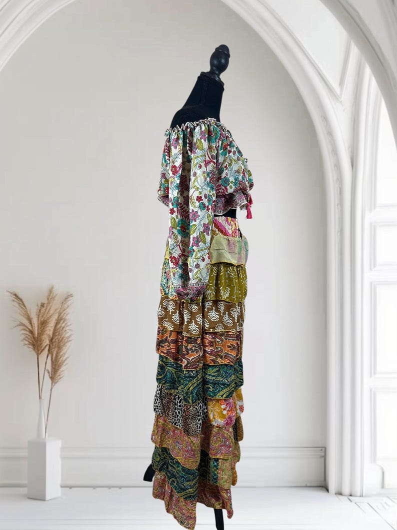 Op de afbeelding: Een kleurrijke, gelaagde maxi-jurk met een off-the-shoulder top. De top heeft een bloemenprint met gefranjerde mouwen en afwerking. De rok is samengesteld uit verschillende stoffen met patronen in tinten bruin, groen, oranje en roze. De jurk wordt getoond op een zwarte mannequin.