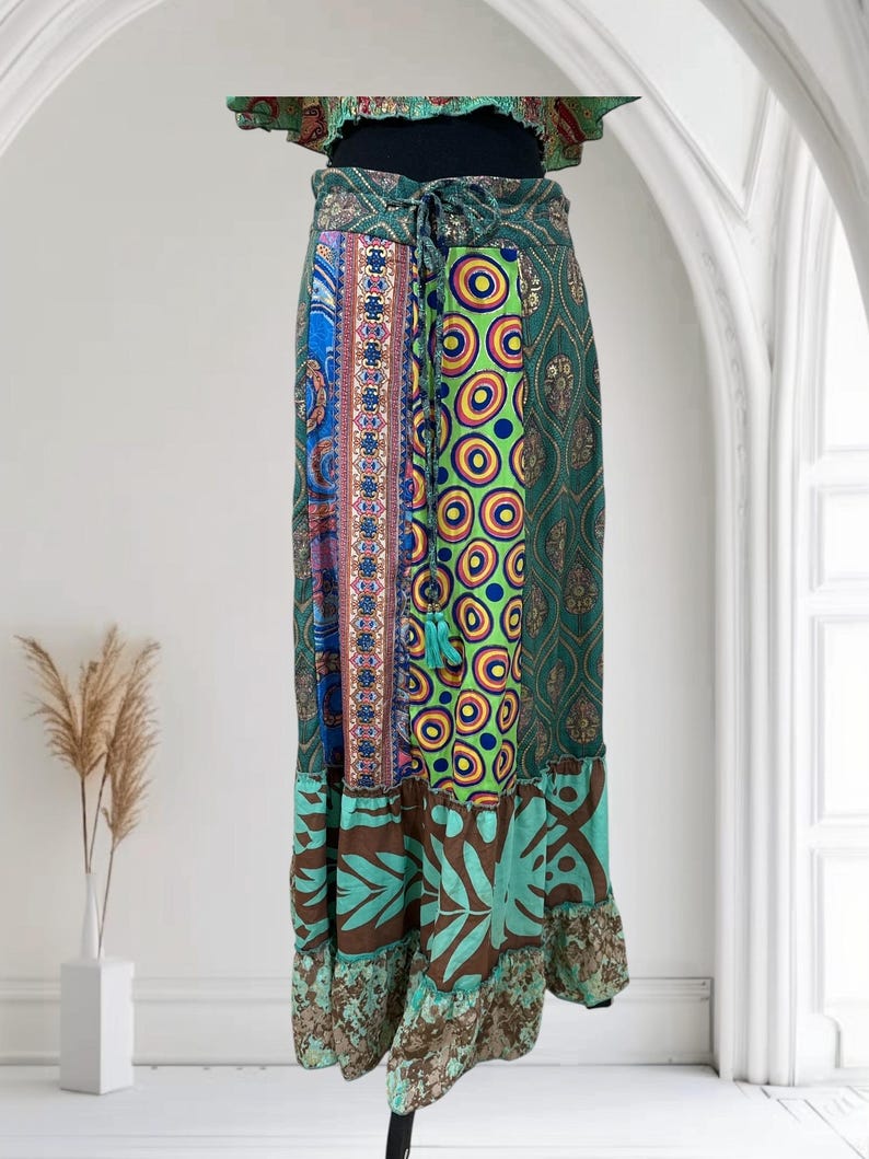 May include: A long, patchwork skirt with a variety of patterns and colors. The skirt features a mix of blue, green, brown, and patterned fabrics. It has a drawstring waist and a ruffled hemline. The skirt is displayed against a white background.