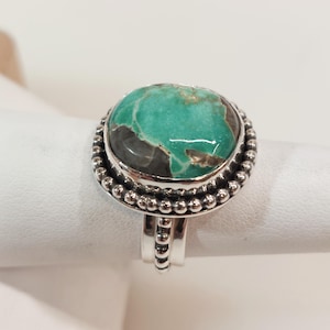May include: A silver ring featuring a large, round turquoise stone with dark gray and brown inclusions. The stone is set in a beaded bezel, and the band has a simple, elegant design. The ring is displayed on a white surface.