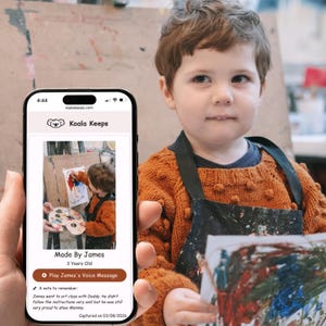 May include: A young child wearing an orange sweater and a black apron holds a painting. A phone displays a Koala Keeps app screen with the text "Made By James, 3 Years Old." The app shows a photo of the child painting.