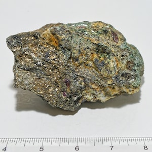May include: A close-up of a rough, gray, green, and gold rock with red crystals. The rock is about 6 inches long and is sitting on a white surface with a ruler.