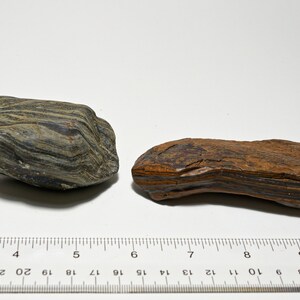 May include: Two pieces of banded iron formation, one is dark gray with black stripes and the other is brown with black stripes. The rocks are about 5 inches long and are sitting on a white surface with a ruler for scale.