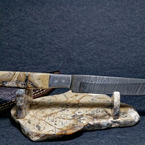 May include: A handmade knife with a patterned steel blade and a handle made of fossilized stone. The knife is displayed on a stand made of the same material.