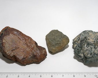 May include: Three rocks of varying sizes and colors are displayed against a white background. The largest rock is reddish-brown, the middle one is gray-green, and the smallest is a mix of gray and green.
