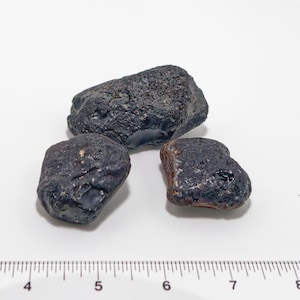 May include: Three dark, irregularly shaped stones with a rough texture are arranged on a white surface. The stones vary in size and have a dark, almost black color, with some areas showing a hint of brown.