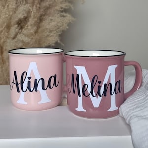 May include: Two personalized pink enamel mugs. One is light pink with the name "Alina" and a white "A". The other is a darker pink with the name "Melina" and a white "M". Both mugs have black rims.