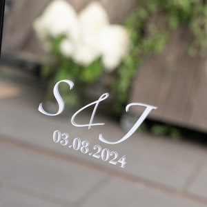 Könnte beinhalten: Eine reflektierende Oberfläche mit den Initialen "S & J" und dem Datum "03.08.2024" in zarter, weißer Schrift. Der unscharfe Hintergrund zeigt Grün und weiße Blumen, was auf eine Hochzeit oder ein besonderes Ereignis hindeutet.